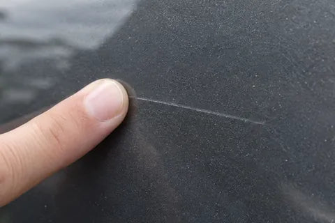 Ein Fingernagel fährt quer über einen feinen Kratzer im Klarlack einer grauen Autotür.